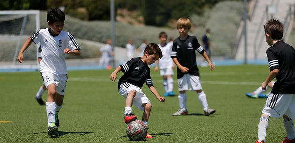 Kids in Real Madrid uniforms playing soccer on a sunny field, with one child controlling the ball while others run nearby.