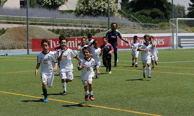 Kids in Real Madrid uniforms running together during a soccer training session on a sunny field, with a coach following behind.