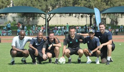 A group of six Real Madrid Foundation coaches kneeling together on a soccer field, smiling at the camera during a youth training session.