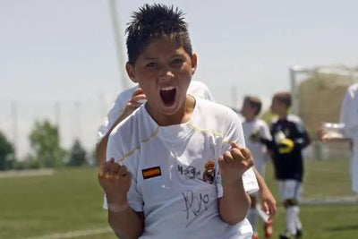 Child in a soccer wearing a Real Madrid uniform celebrating on a field with other players in the background.
